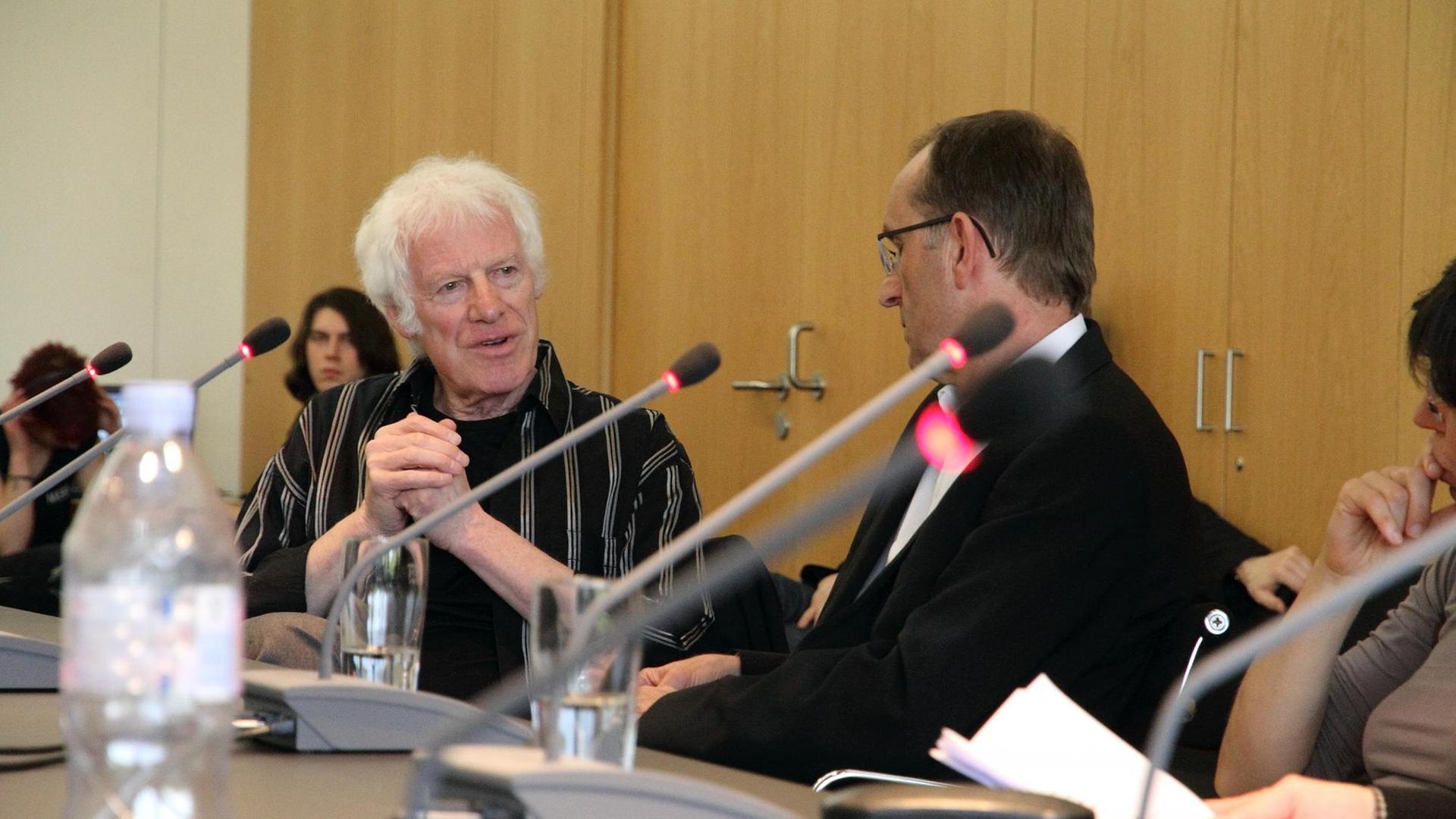 Ein alter Mann mit weißen Haaren sitzt an einem langen Tisch und spricht. Es ist der Komponist Rolf Riehm. Das Foto ist beim Forum Neuer Musik entstanden. Ein alter Mann mit weißen Haaren sitzt an einem langen Tisch und spricht. Es ist der Komponist Rolf Riehm. Das Foto ist beim Forum Neuer Musik entstanden.