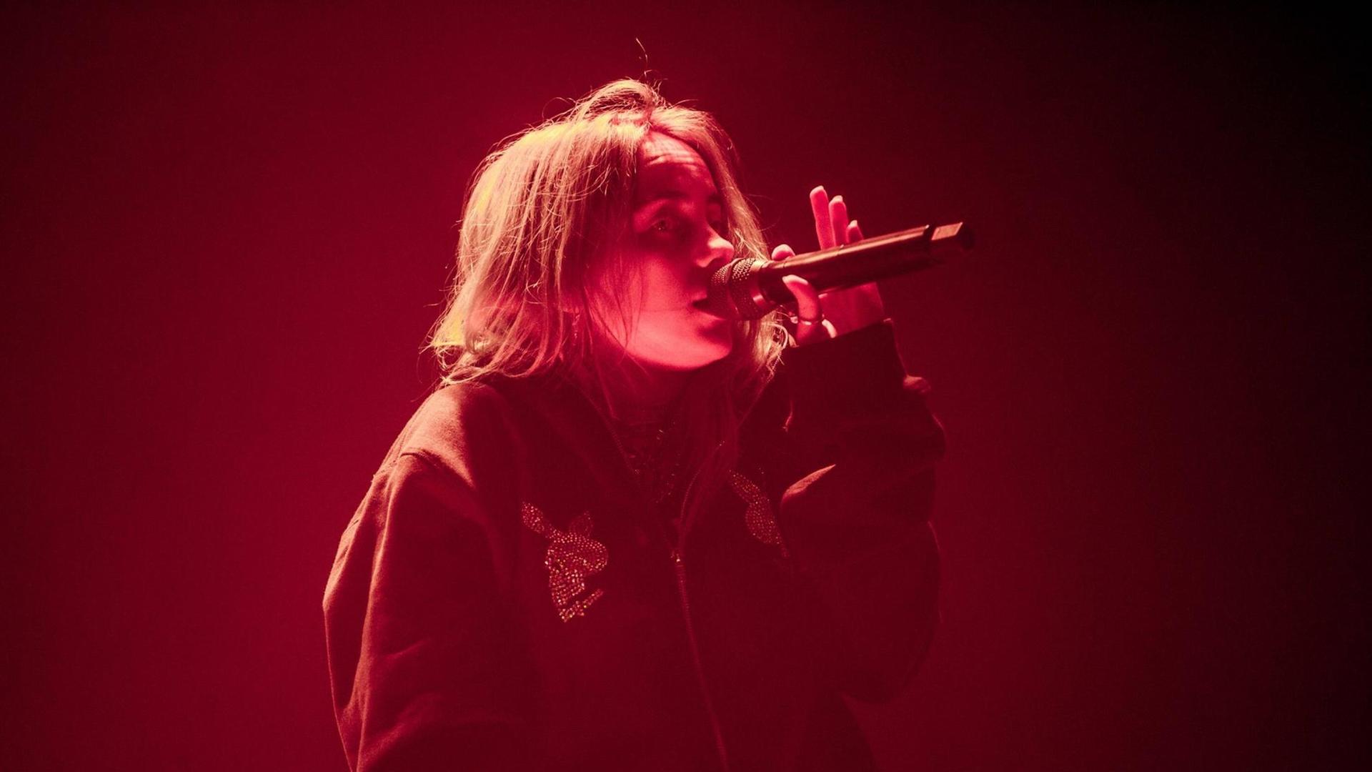 Die junge Sängerin Billie Eilish steht in rotem Licht auf der Bühne und singt mit einem Mikrofon in der linken Hand.