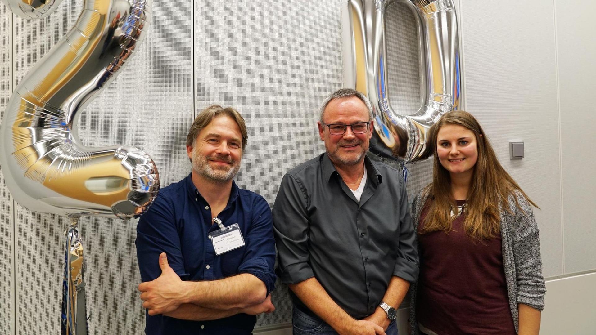 Oliver Janoschka, Prof. Jürgen Handke, Louisa Oesterle (v.l.) stehen im Studio. Im Hintergrund schweben silberfarbene Luftballons, welche die Zahl zwanzig bilden.