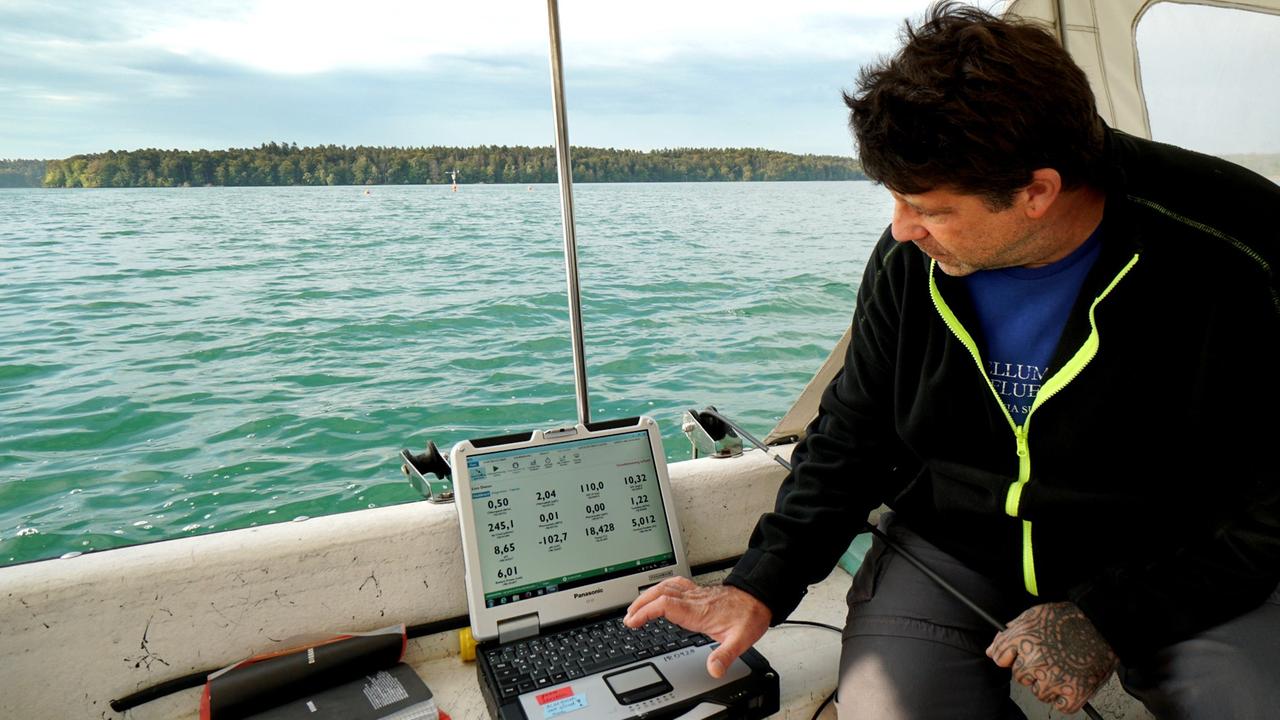 Ein Mann sitzt in einem Boot auf einem See. Vor ihm steht ein Computer. 