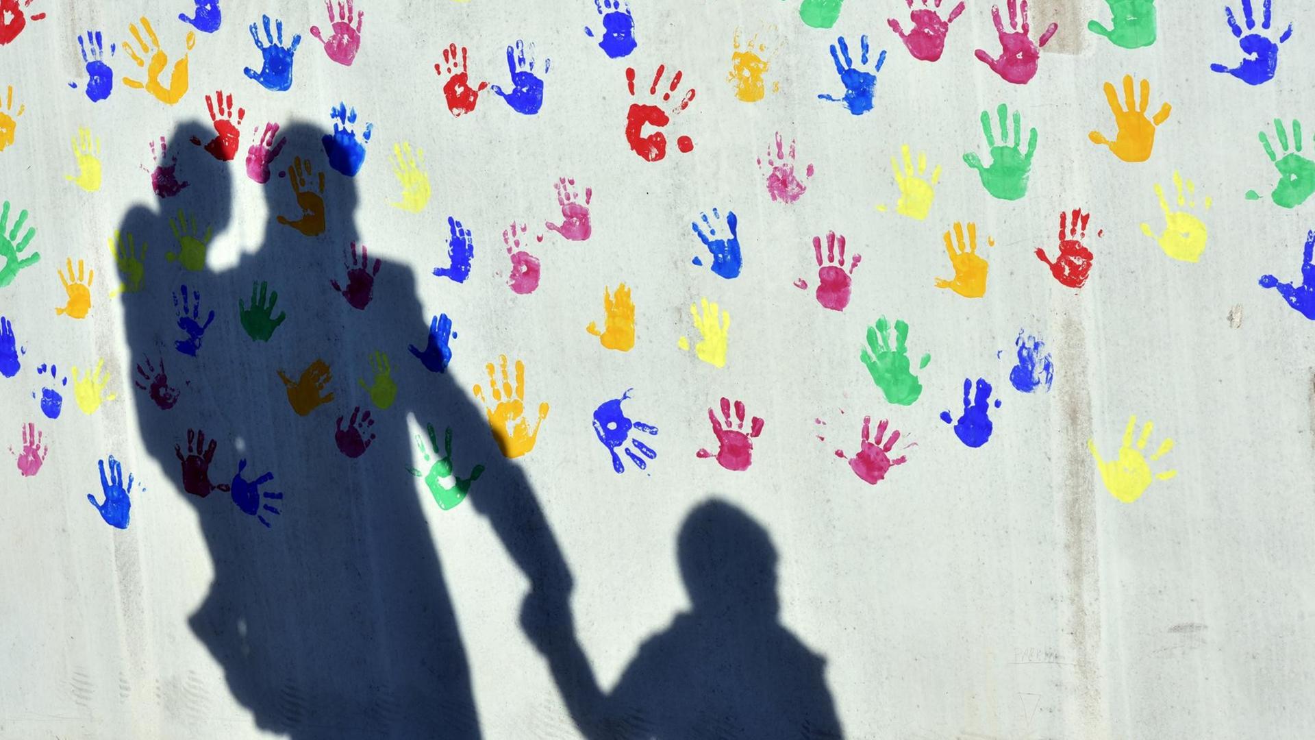 Ein Mann mit einem Kind auf dem Arm und einem an der Hand wirft einen Schatten auf eine mit bunten Handabdrücken bemalte Wand einer Kindertagesstätte.