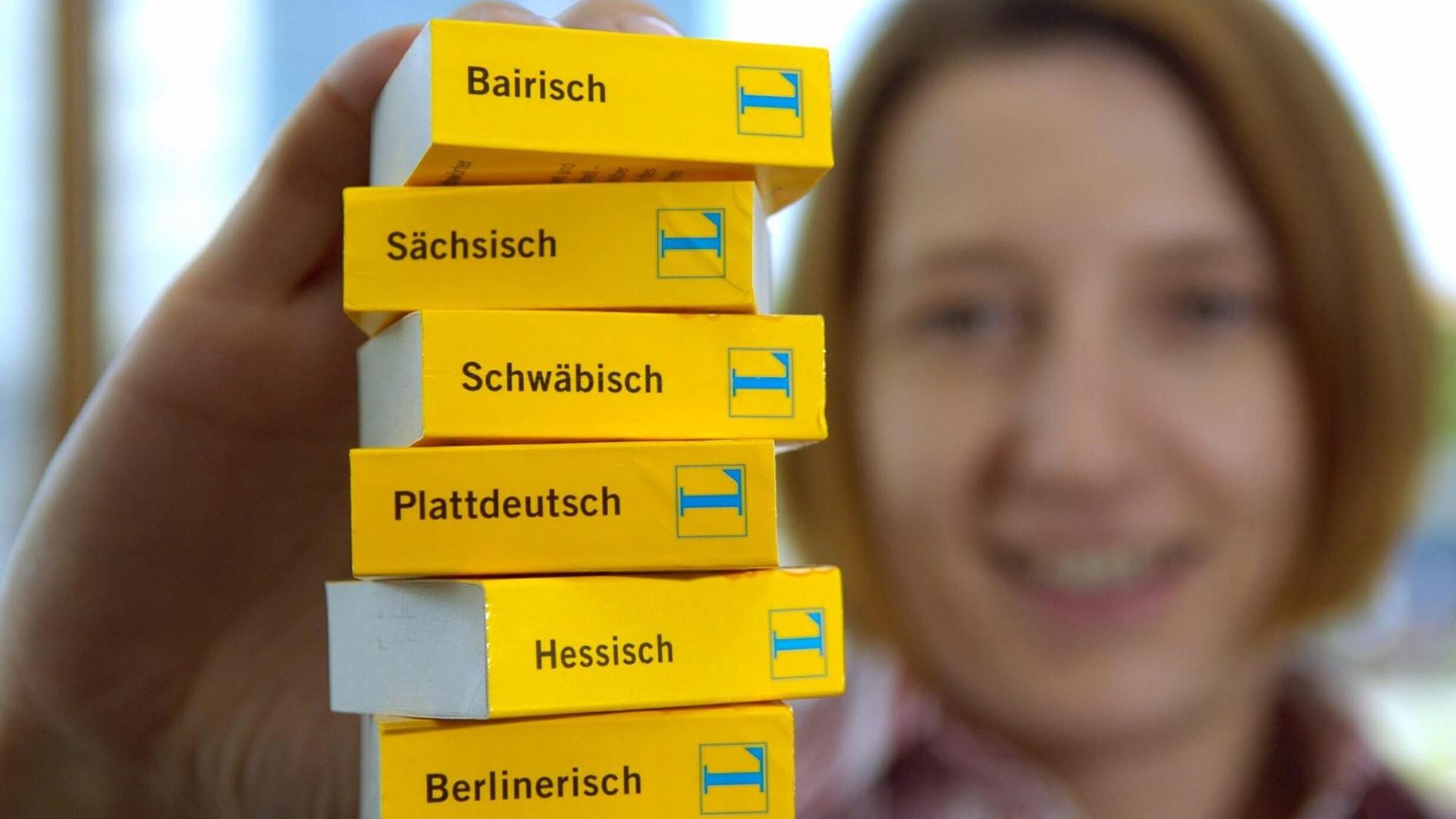 Eine Frau hält einen Stapel Mini-Wörterbücher von Langenscheidt in den Ausgaben Hessisch, Plattdeutsch, Sächsisch, Schwäbisch, Berlinerisch und Bairisch in die Kamera