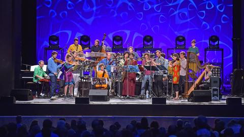 Ein bunt gemischtes Orchester spielt auf einer Bühne mit blau-animiertem Hintergrund vor Publikum. Die meisten Musiker stehen. Unter den Instrumenten: Harfe, Fagott, Baritonsaxofon, Flöte, Cello und Geigen.