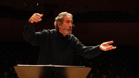 Jordi Savall dirigiert ohne Taktstock mit ausgebreiteten Händen, er trägt ein schwarzes Hemd mit Stehkragen, Bart und Brille, und vor ihm steht ein Notenpult