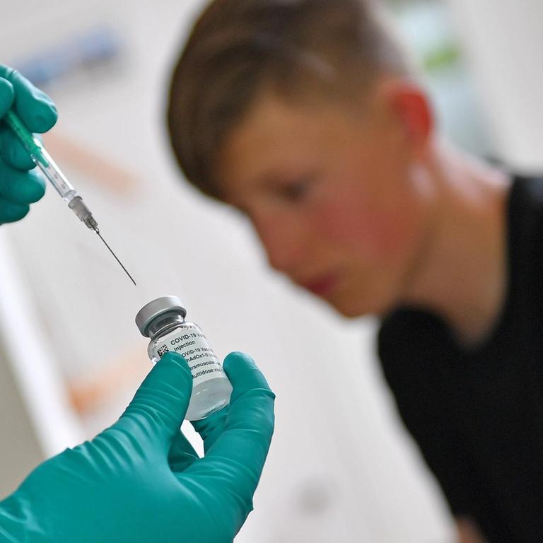 Ein Jugendlicher kurz vor einer Impfung, im Vordergrund sind Hände in Handschuhen zu sehen, die eine Spritze halten. 
