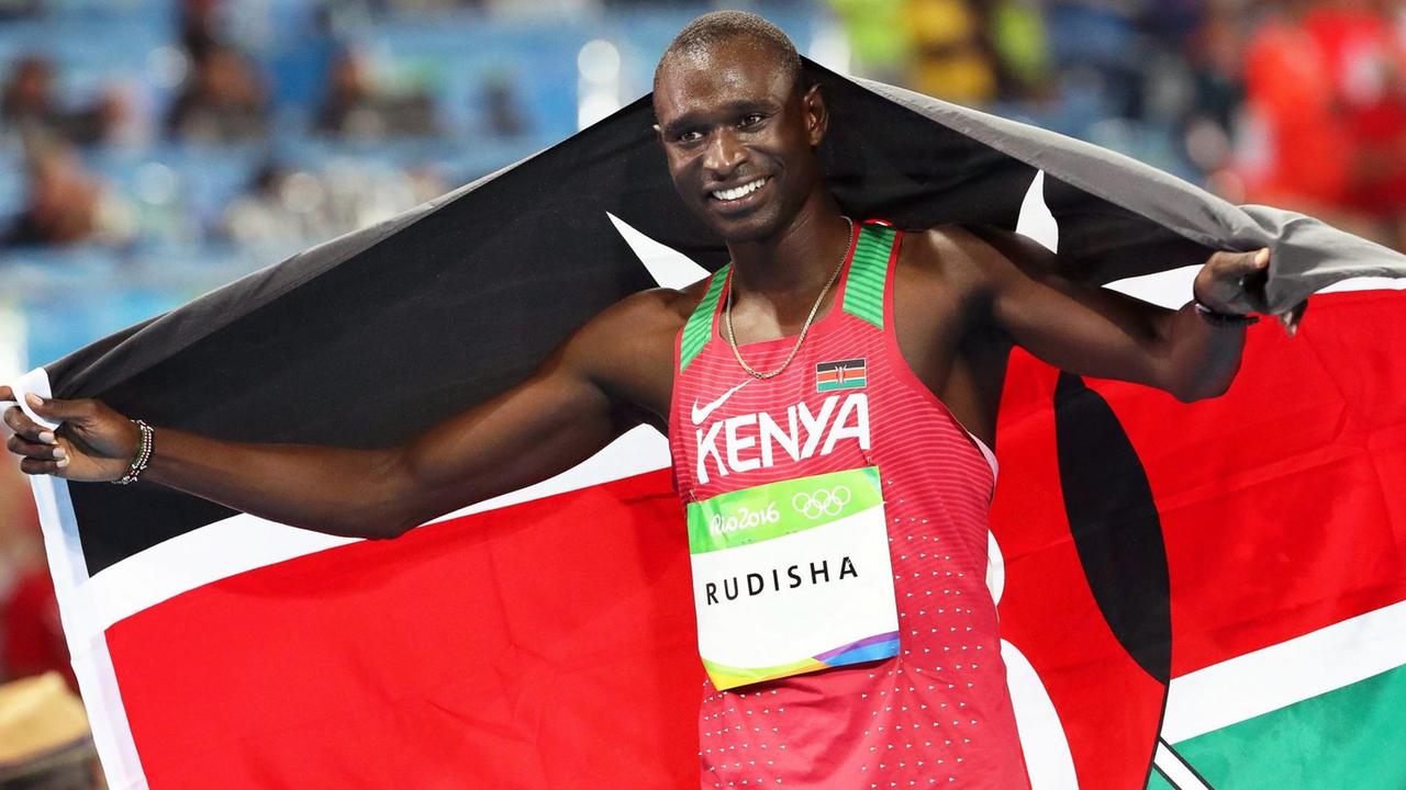 Der Kenianer David Lekuta Rudisha hält lächelnd die kenianische Nationalflagge hinter seinen Rücken.