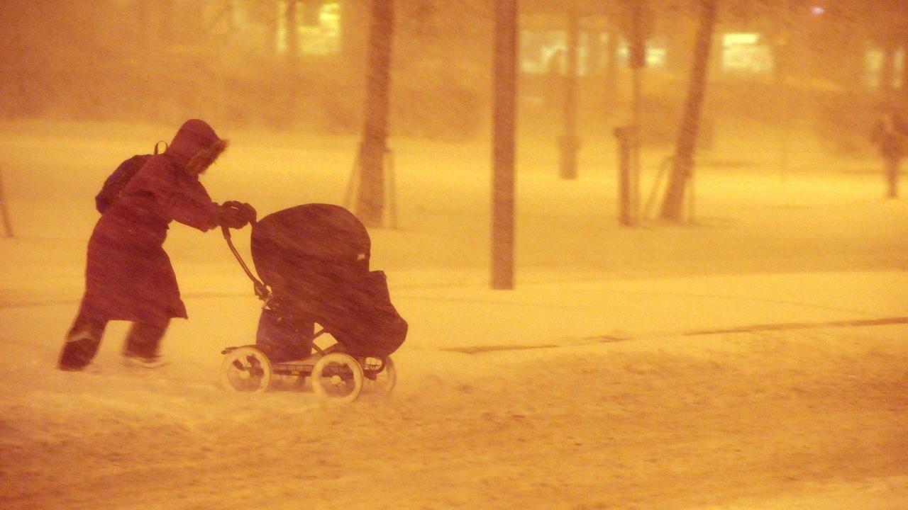 Eine Person im Wintermantel schiebt einen Kinderwagen durch einen Schneesturm.