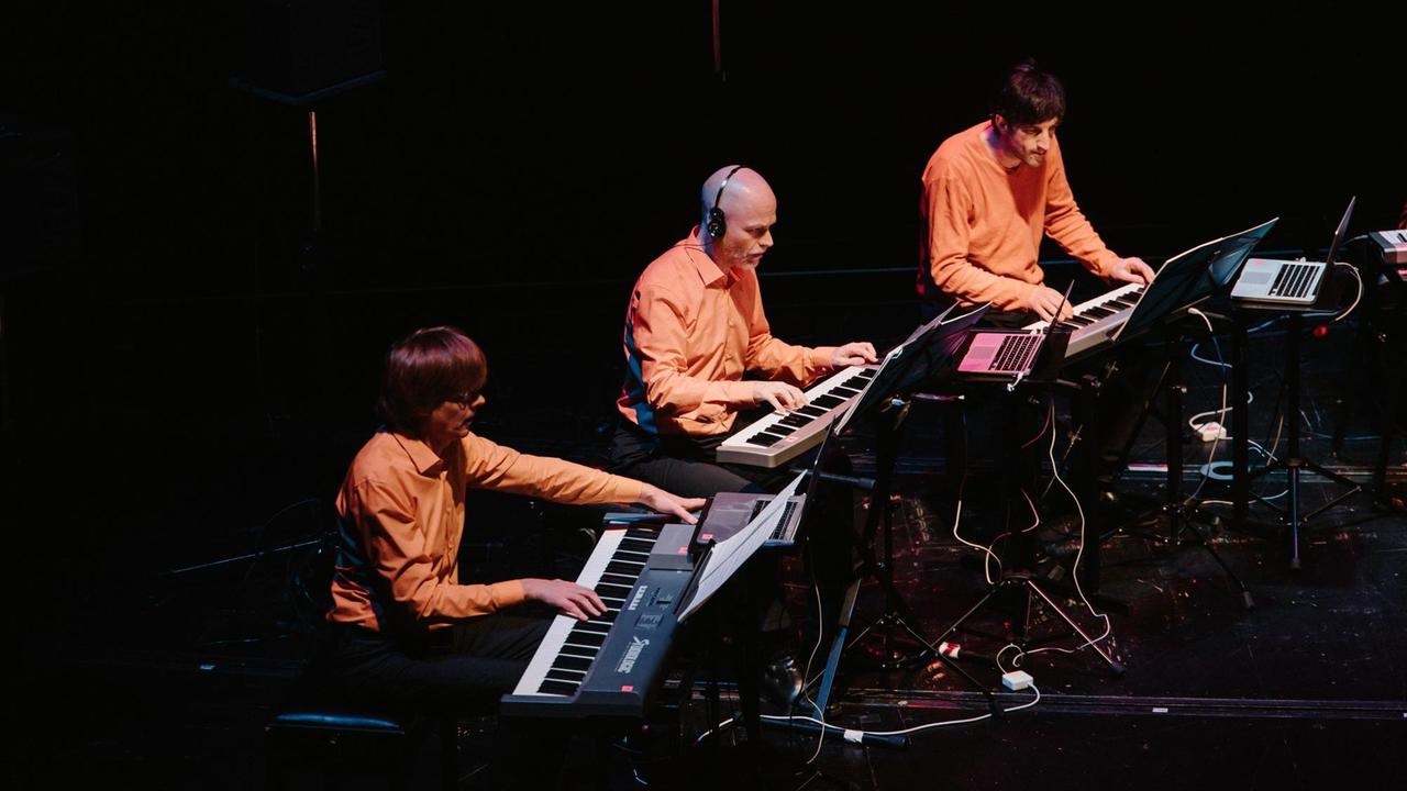 Drei der Musiker sitzen im orangefarbenen Hemd an elektronsichen Tastaturen
