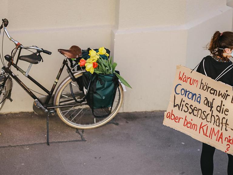 Eine junge Frau mit Mundschutz trägt ein Papschild auf dem Rücken auf dem steht: "WArum hören wir bei Corona auf die Wissenschaft, aber beim Klima nicht?" Im Hintergrund steht ein Fahrrad mit Blumen.