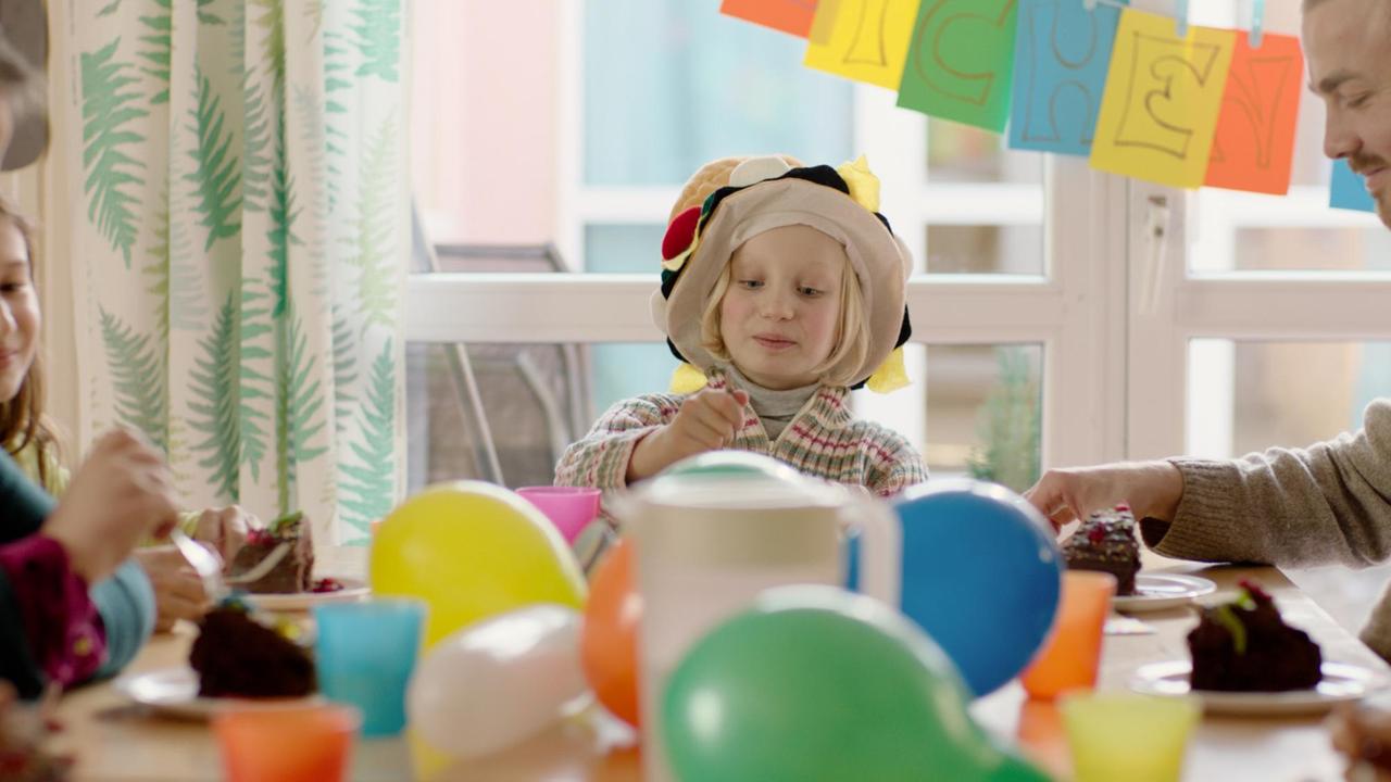Ein kleines blondes Mädchen mit einer bunten Kopfbedeckung. Es sitzt an einem Kaffeetisch der für eine Geburtstagsfeier mit Luftballons geschmückt ist. Über ihr hängt eine bunte Girlande.