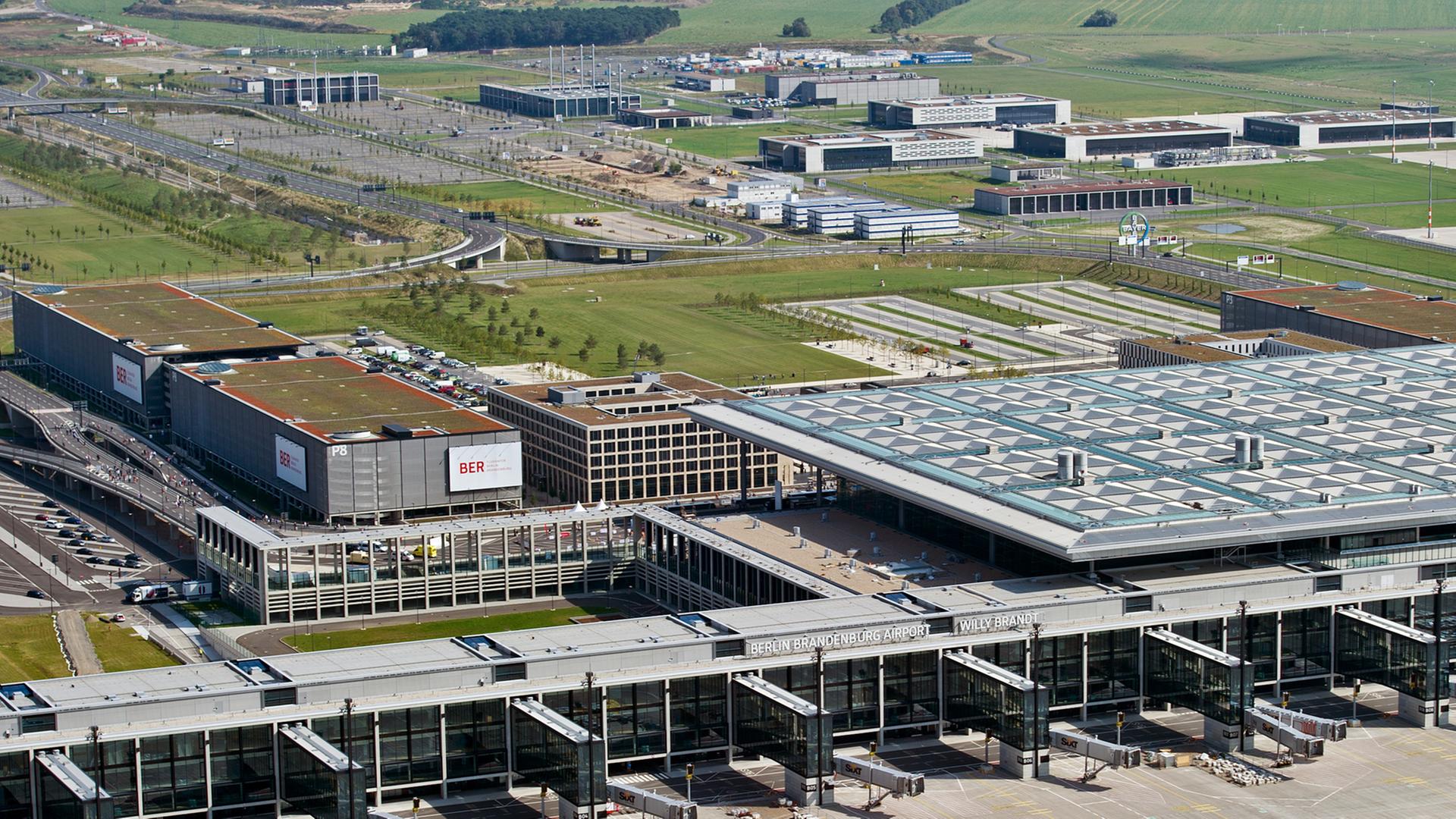 Luftbild eines Terminals des Flughafens mit Vorfeld, Gebäude und Gewerbegebäuden im Hintergrund.