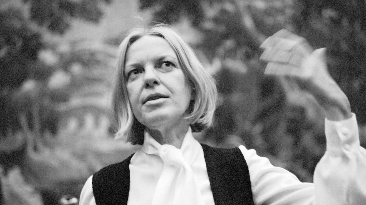 Ein Schwarzweiß-Foto zeigt die österreichische Lyrikerin Ingeborg Bachmann 1972 nach der Verleihung des Wildganspreises in weißem Hemd und schwarzer Weste auf einer Bank sitzend, die linke Hand erhoben gestikulierend, den Blick leicht lächelnd nach oben gerichtet.