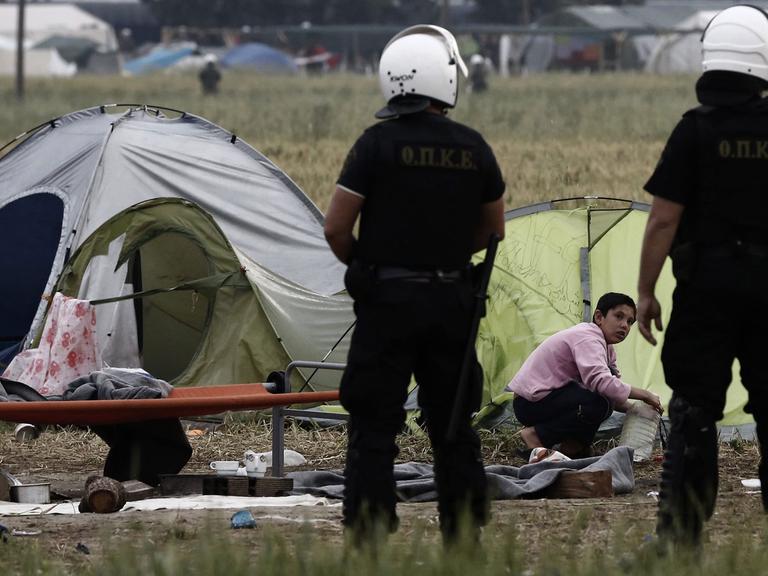 Zwei griechische Polizisten stehen bei der Räumung des des Flüchtlingslagers in Idomeni am 24.05.2016 vor Zelten, vor denen ein Flüchtlingsjunge kauert.