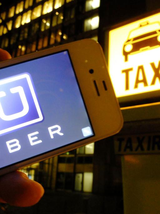 Es ist Nacht in einer Stadt: Eine Hand hält ein Smartphone mit dem Uber-Logo in die Kamera; im Hintergrund ist eine Taxirufsäule mit dem beleuchteten "Taxi"-Logo zu sehen.