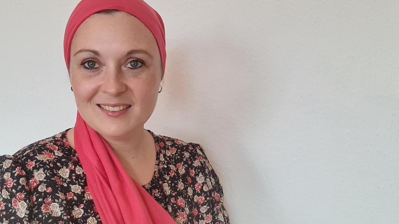 Eine Frau mit rotem Kopftuch steht vor einer weißen Wand und lächelt in die Kamera