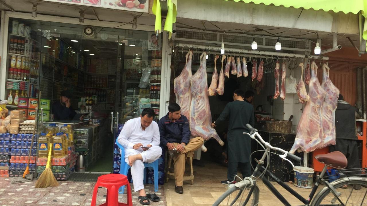 Blick auf kleine Läden in Kabul. In der Auslage hängen große Stücke Fleisch an Haken. Vor dem benachbarten Geschäft sind Getränkekisten aufgebaut. 