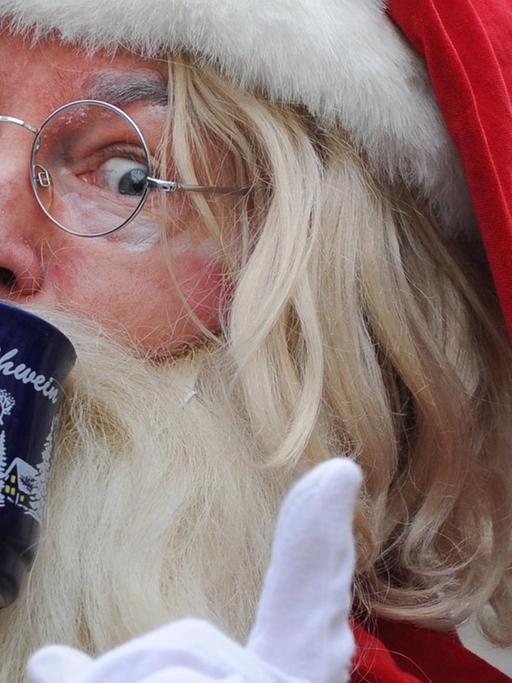 Ein als Weihnachtsmann verkleideter Mann trinkt Glühwein und hebt dabei den Zeigefinger.