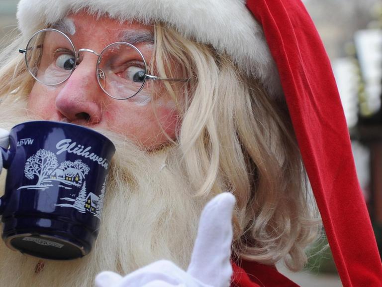 Ein als Weihnachtsmann verkleideter Mann trinkt Glühwein und hebt dabei den Zeigefinger.