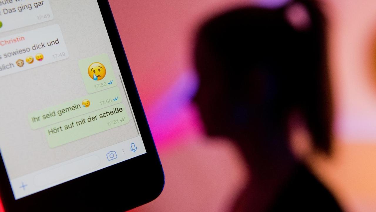 Die Silhouette eines junges Mädchens im Hintergrund, davor ist auf dem Display eines Smartphones ein fiktiver Chatverlauf beim Messenger "WhatsApp" zu sehen.