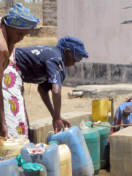 Dorfbewohnerinnen füllen am Brunnen Wasser ab, im Hintergrund die Tanks, in denen das aufgefangene und gefilterte Regenwasser gelagert wird.