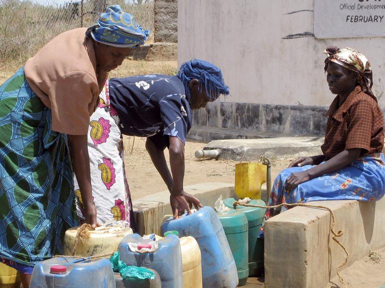 Dorfbewohnerinnen füllen am Brunnen Wasser ab, im Hintergrund die Tanks, in denen das aufgefangene und gefilterte Regenwasser gelagert wird.