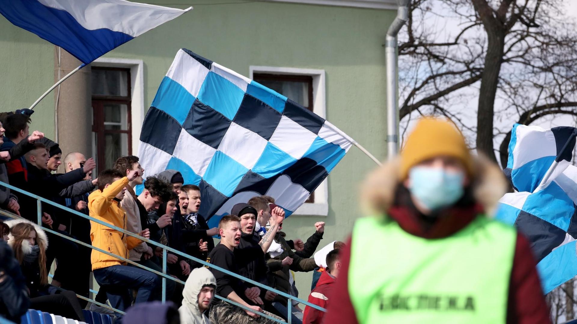 Fans im Fanblock beim Fußballspiel zwischen FC Slutsk und Slavia Mazyr, im Vordergrund ein Ordner mit einem Mundschutz