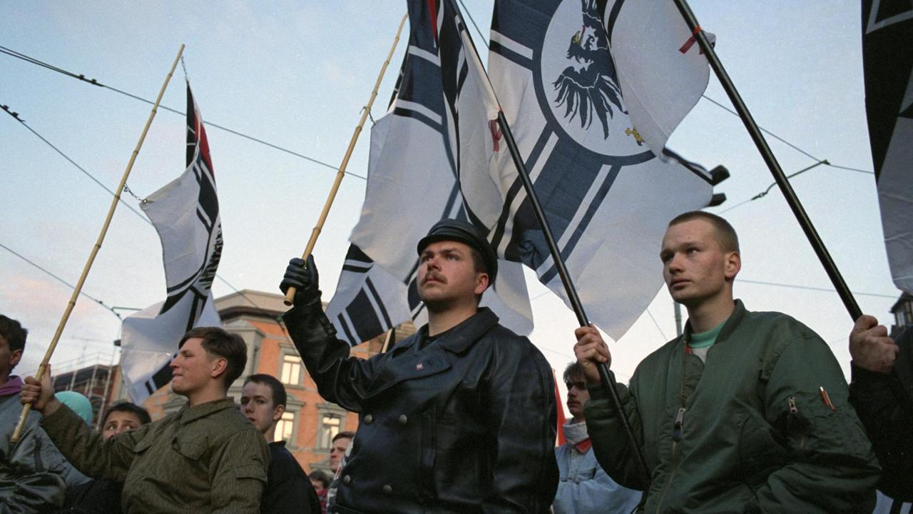 Neonazis stehen mit Reichskriegsflaggen in der Hand zusammen. Ihr Blick ist ernst und entschlossen.