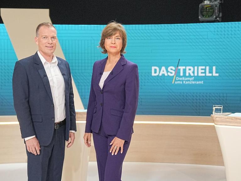 Die Moderatoren Oliver Köhr und Maybrit Illner stehen im Fernsehstudio.