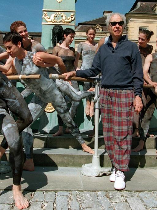 Merce Cunningham steht auf dem Platz vor dem Ludwigsburger Schloss, hinter und neben ihm sein Ensemble in dunklen, engen Kostümen in Tanzposen.