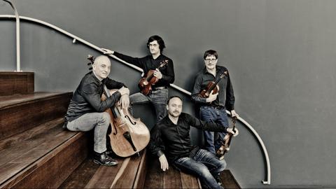 Ein Cellist und ein Geiger sitzen mit ihren Instrumenten auf einer hölzernen Treppe. Dahinter stehen ein Bratschist und ein Geiger, die sich an die weiße Treppenstange anlehnen.