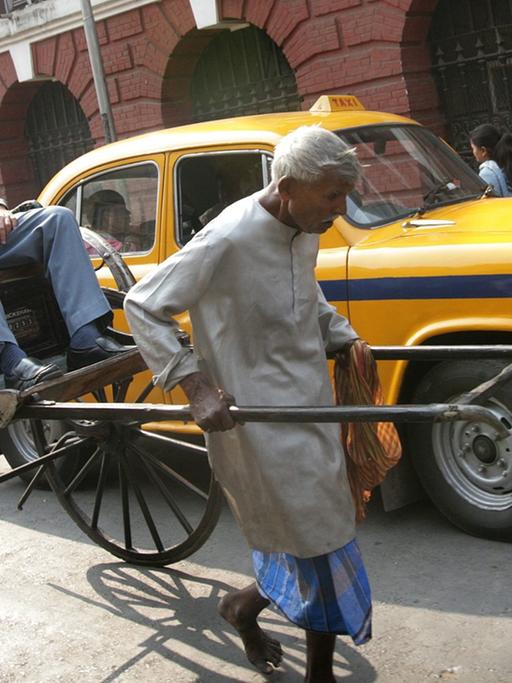 Ein älterer Rikscha-Fahrer zieht seine Rikscha mit einem Fahrgast durch eine Straße Kalkuttas, dahinter steht ein gelbes Taxi.