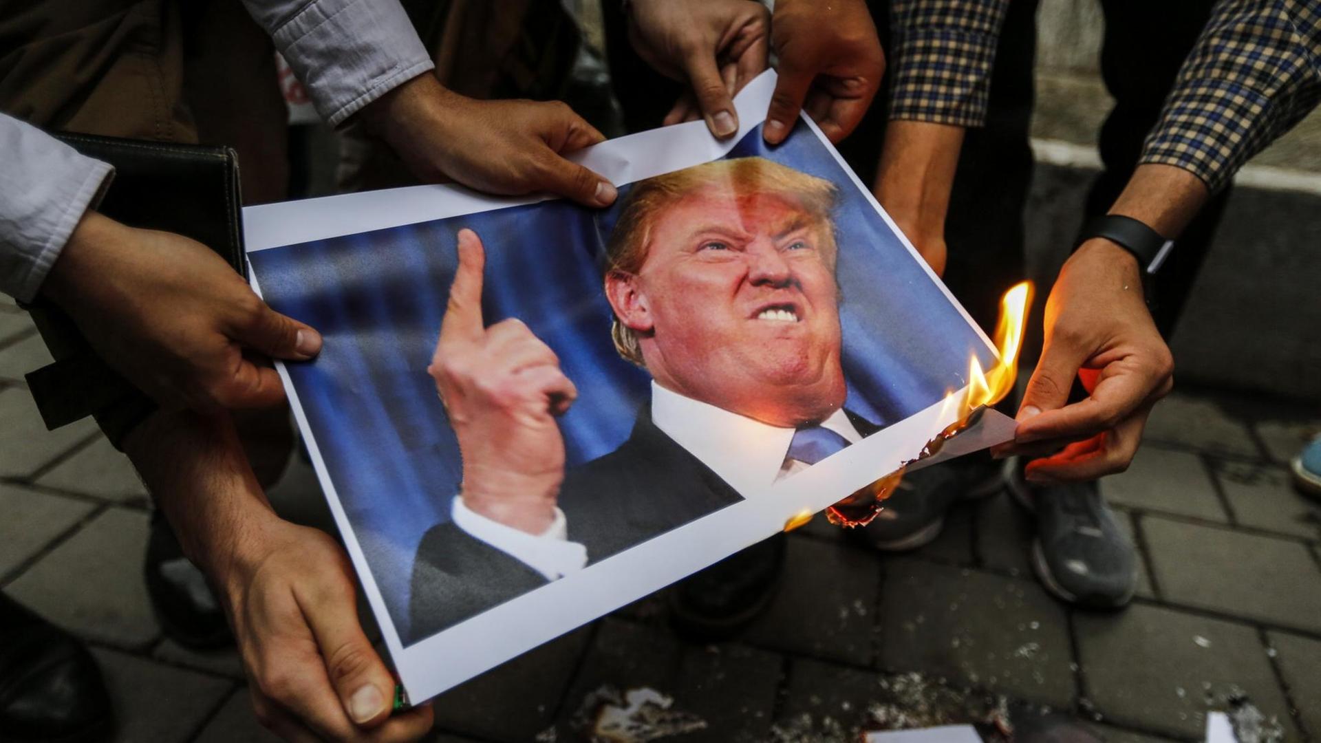 Man sieht Hände. Sie halten ein Bild von dem Präsidenten von dem Land USA fest. Eine Hand hält ein brennendes Feuerzeug an das Papier.