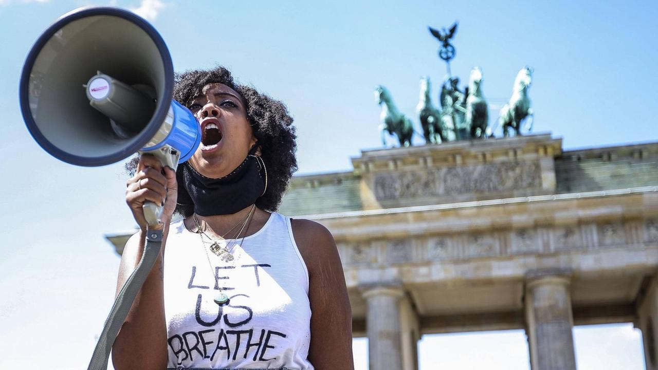 Eine schwarze Frau mit einem beschrifteten weißen Shirt steht vor dem Brandenburger Tor, sie ruft in ein Megafon.