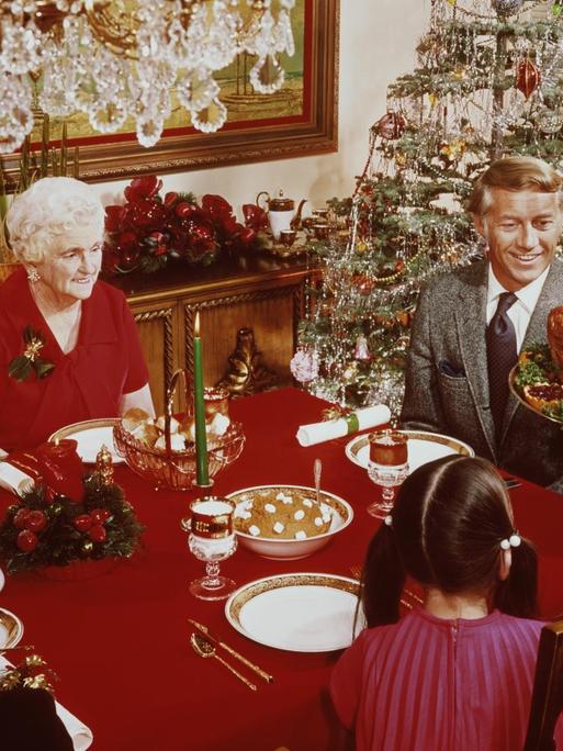 Eine Aufnahe von 1965 zeigt eine Frau, die einen großen Truthahn zum Weihnachtsessen auf den Tisch bringt. Dort sitzen Großeltern und Kinder am gedeckten Tisch. Im Hintergrund ist ein geschmückter Weihnachtsbaum zu sehen.