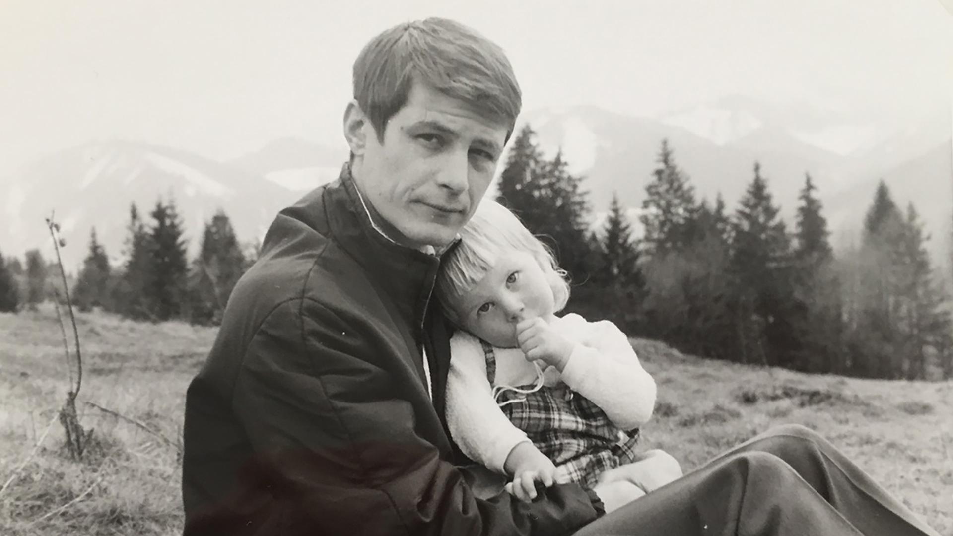 Vater und Tochter sitzen in den Bergen, die Tochter schmiegt sich an ihren Vater und nuckelt am Daumen.