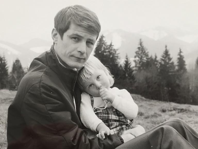 Vater und Tochter sitzen in den Bergen, die Tochter schmiegt sich an ihren Vater und nuckelt am Daumen.
