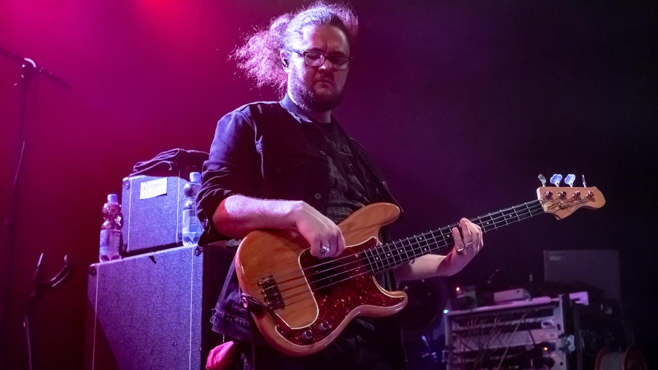 Ein Mann mit Zopf und Brille steht auf einer Bühne und hält eine Bass-Gitarre in der Hand.
