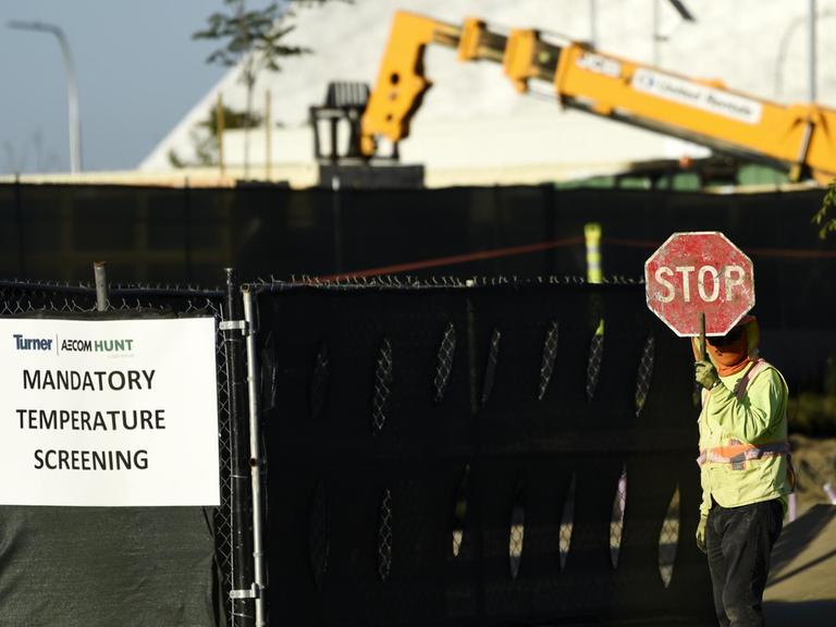 Ein Arbeiter steht an einem Tor zur Baustelle mit "Stop"-Schild, damit niemand ohne Temperaturmessung auf das Gelände kommt.