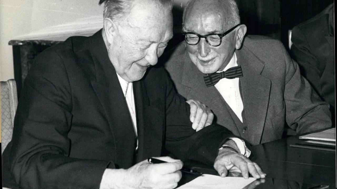 Bundeskanzler Konrad Adenauer sitz zu einer Unterschrift an einem Tisch neben Justizminister Fritz Schaffer. Dieser fasst Adenauer am Unterarm, beide lachen.
