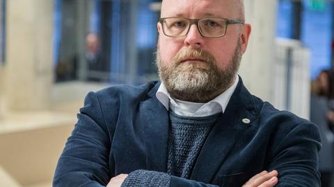 Ein Mann mit Vollbart und verschränkten Armen steht in einem Foyer und schau ernst in die Kamera.