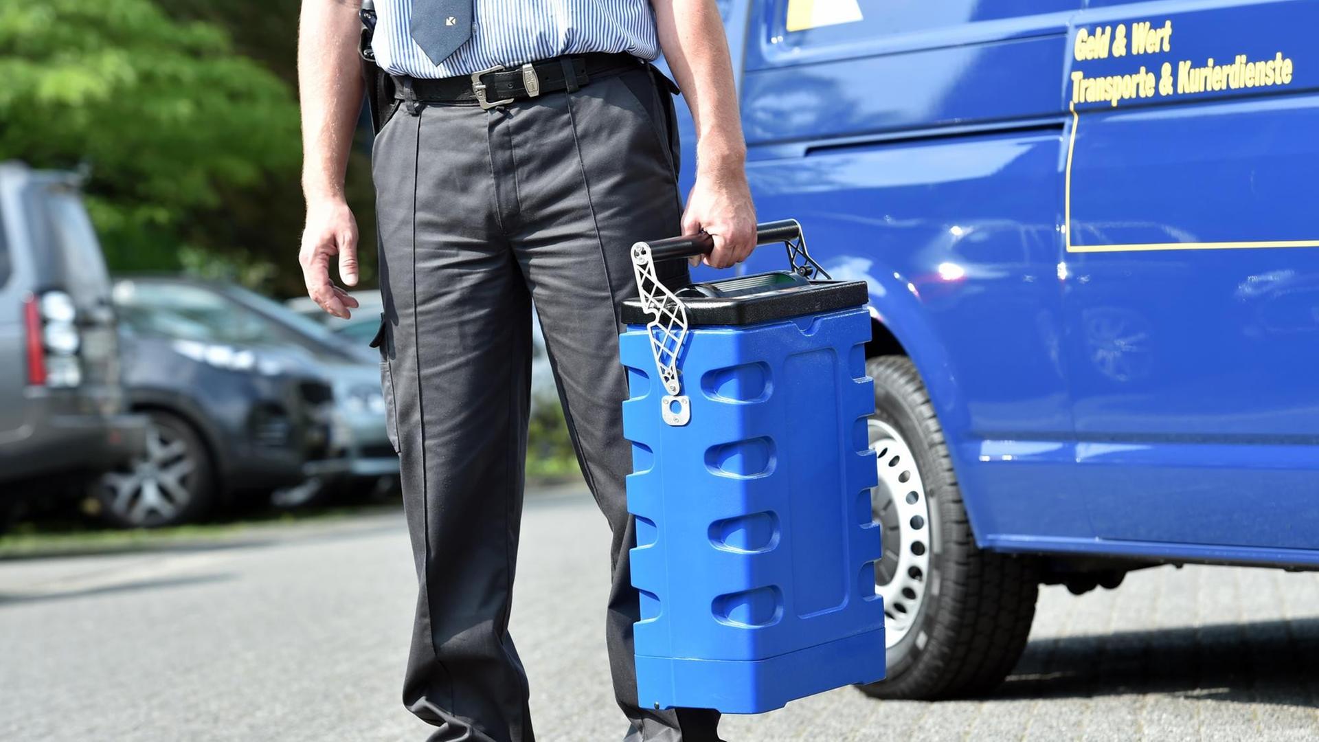 Man sieht nur den Unterkörper des Mannes, der die blaue Geldkassette trägt. Dahinter ein blauer Transporter mit der Aufschrift "Geld & Wert - Transporte und Kurierdienste".