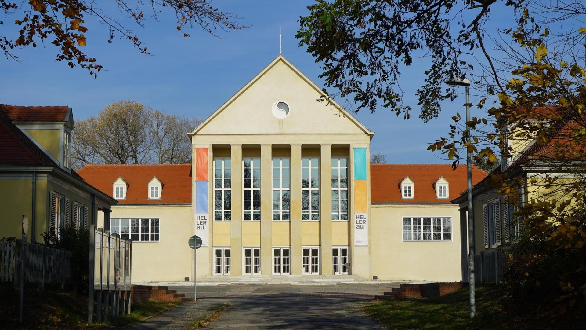 Das Festspielhaus Hellerau ist ein großes Gebäude mit vielen Fenstern an der Front. Die Fassade ist hellgelb, man sieht durch Blätter hindurch auf das Haus.