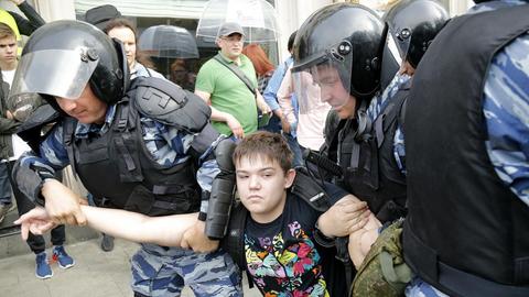 Der Junge im Tiger-Shirt wird mit unbewegter Miene von drei Polizisten in Kampfausrüstung abgeführt. Dahinter stehen weitere Demonstranten.