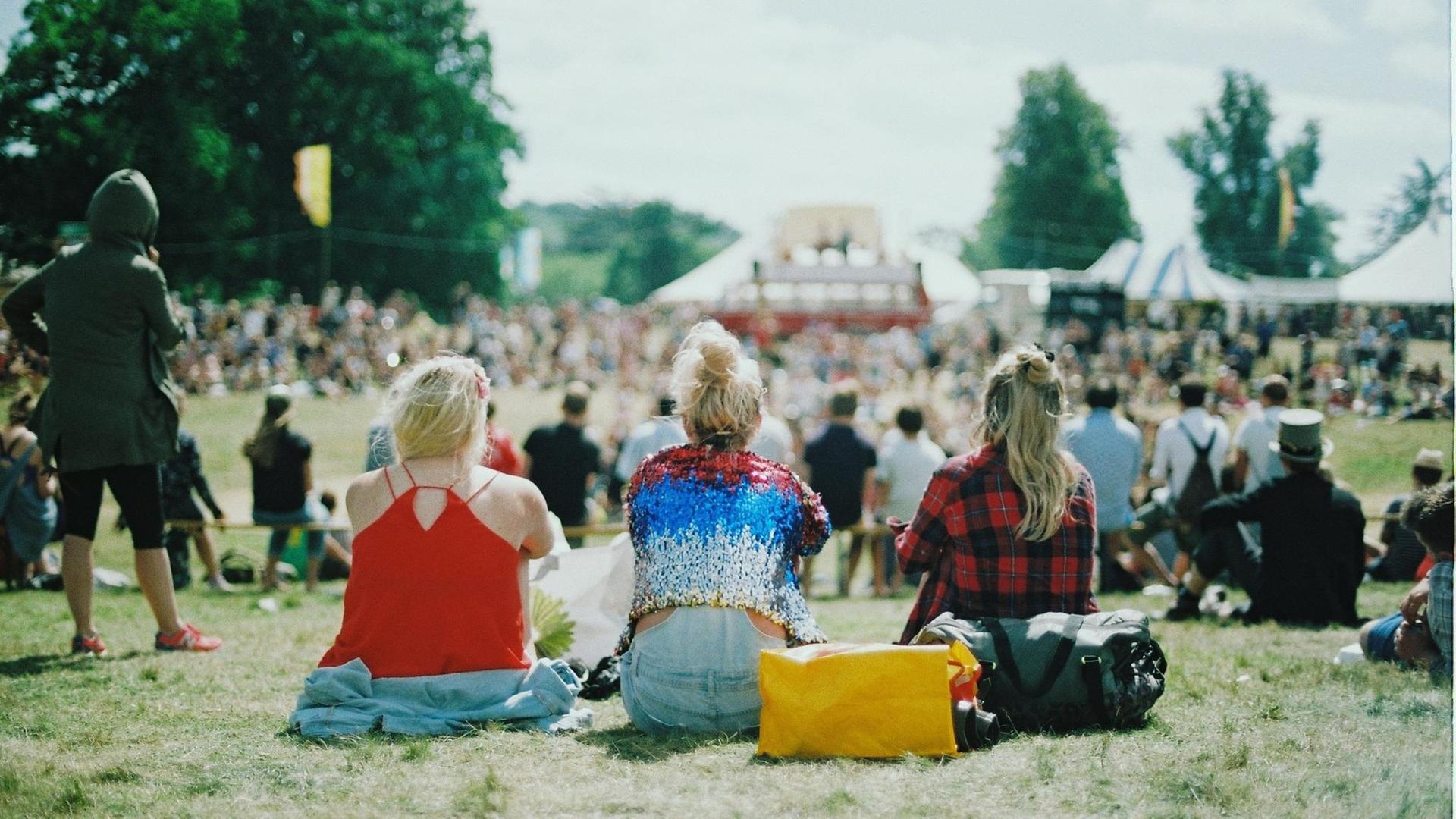 Drei junge Frauen sitzen gemeinsam auf einer Wiese und schauen auf das Festivalgelände.