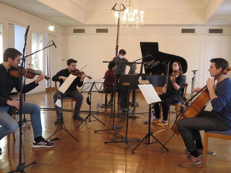 Fünf Musiker an ihren Streichinstrumenten und am Flügel zwischen Mikrofonen in einem Festsaal mit Parkettboden.