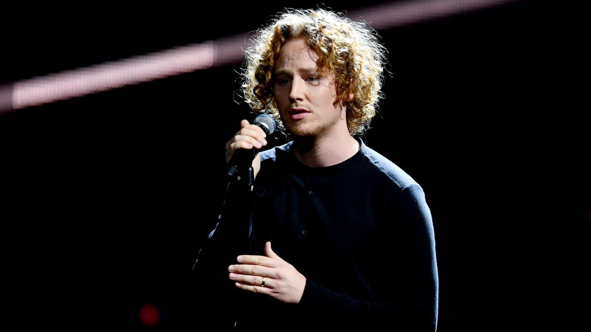 Michael Schulte - gekleidet in schwarz vor schwarzem Hintergrund - spricht auf einer Bühne in ein Mikrofon. Michael Schulte - gekleidet in schwarz vor schwarzem Hintergrund - spricht auf einer Bühne in ein Mikrofon.