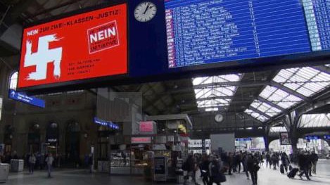 Ein großes Plakat hängt am Züricher Bahnhof neben der Anzeigetafel, darauf ist ein zum Hakenkreuz verschmiertes Schweizer Kreuz zu sehen.