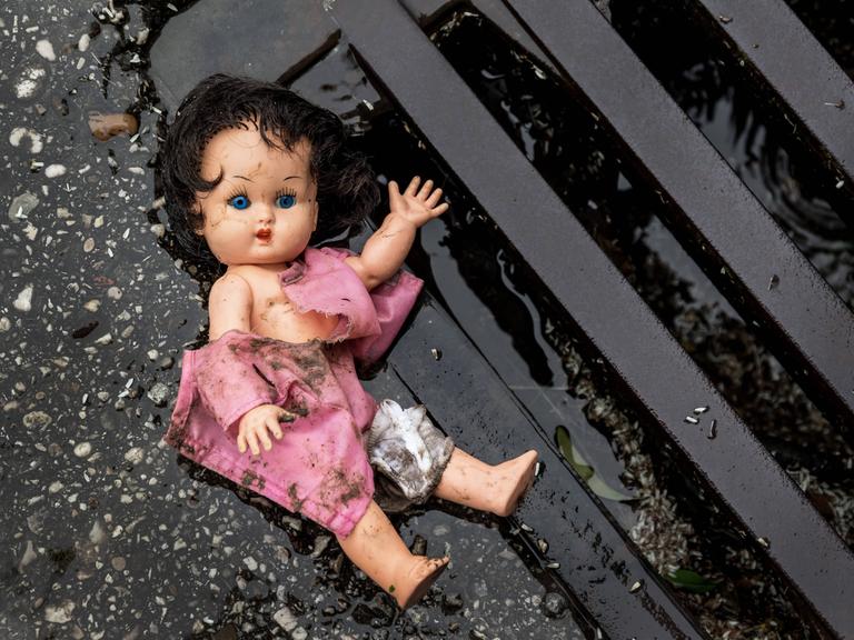 Eine Puppe liegt in dreckiger Bekleidung an einem Straßengully.