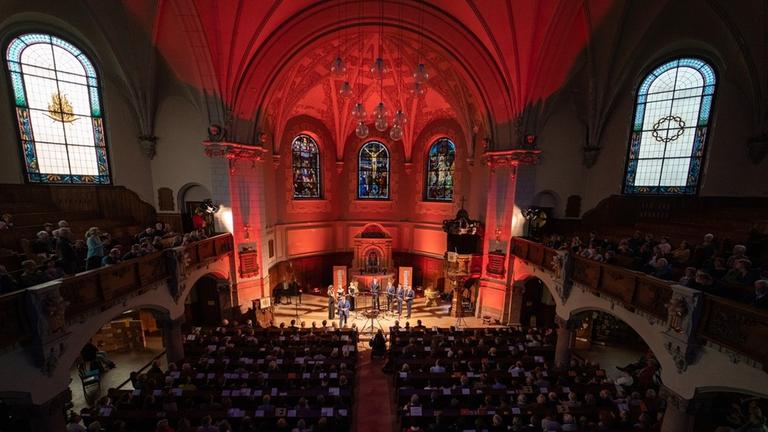 Drei Sängerinnen, fünf Sänger stehen im Halbkreis auf der Bühne. Der Kirchenraum ist orange beleuchtet.