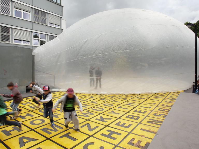 Küchenmonument, Eine Soziale Skulptur vor dem Museum, Berlinische Galerie, 2014. Auf dem Bild sind rennende Kinder auf einem Feld mit lauter Buchstaben zu sehen, dahinter Menschen, eingehüllt in eine riesige Blase.